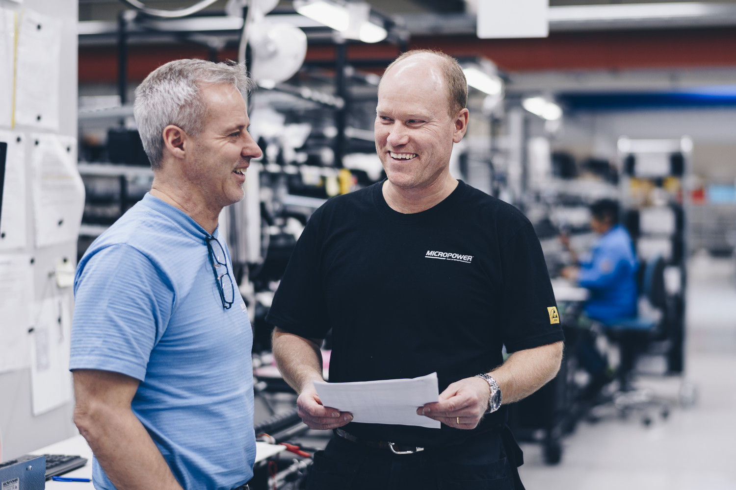 Two male employees at Micropower Group are talking and smiling in the battery factory.