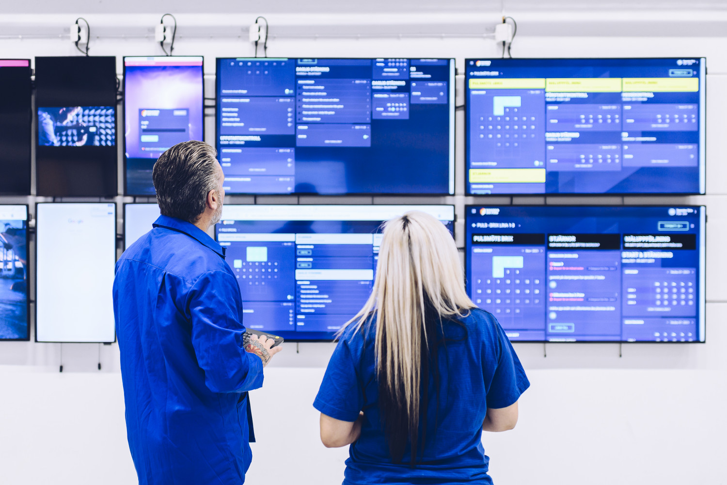 A man and a woman in blue overalls are looking at several large computer screens in a clean industrial environment.